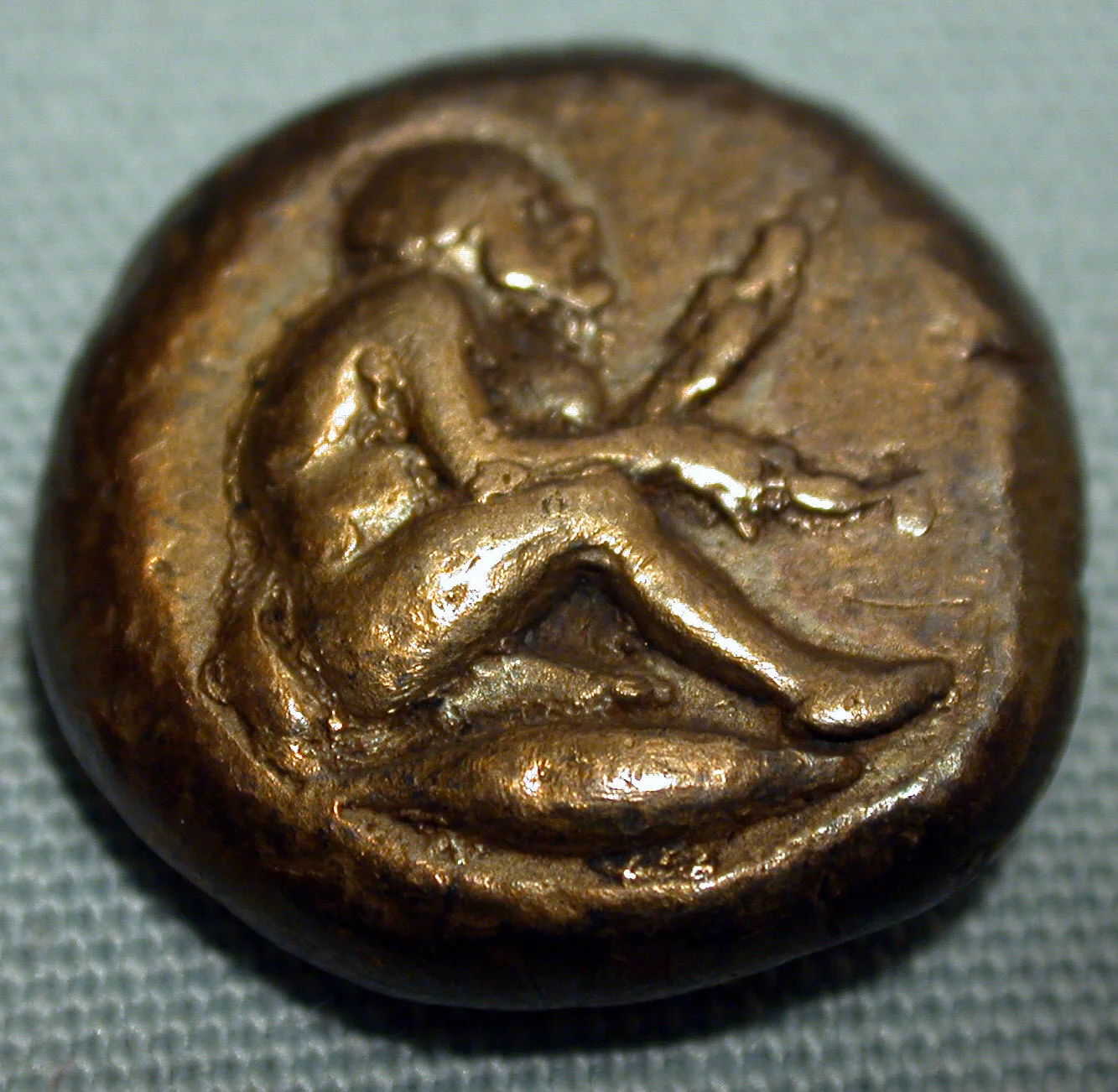 A thick globular ancient coin showing a squatting figure, the entire surface copper-brown rather than silver-white