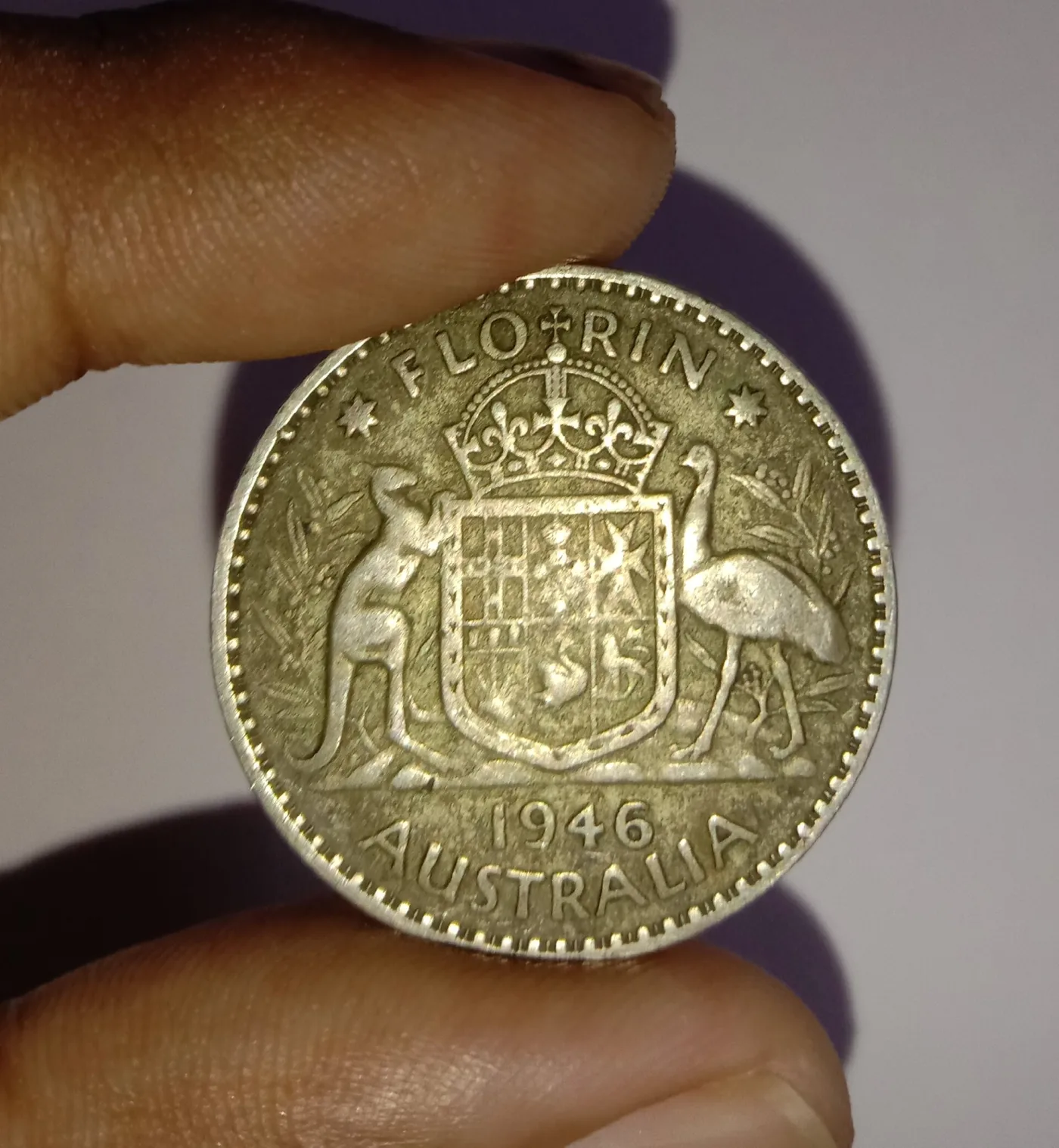 Reverse of an Australian florin dated 1946 held between finger and thumb, a crowned shield divided into six compartments supported by a kangaroo and an emu