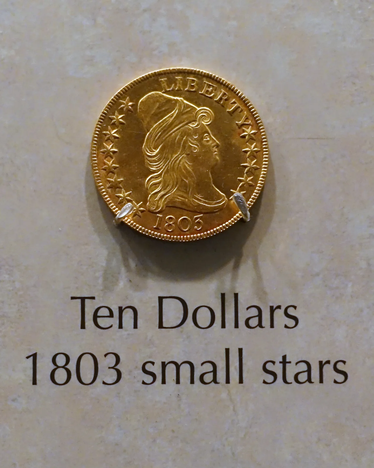 A gold eagle dated 1803 mounted in a museum case above a printed label reading Ten Dollars, 1803 small stars