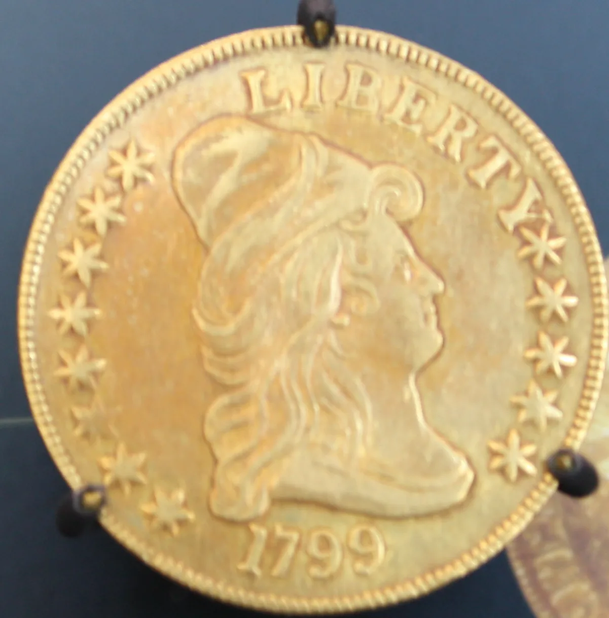 Obverse of a gold eagle dated 1799 held in a display stand, with large boldly punched stars around Liberty's capped bust