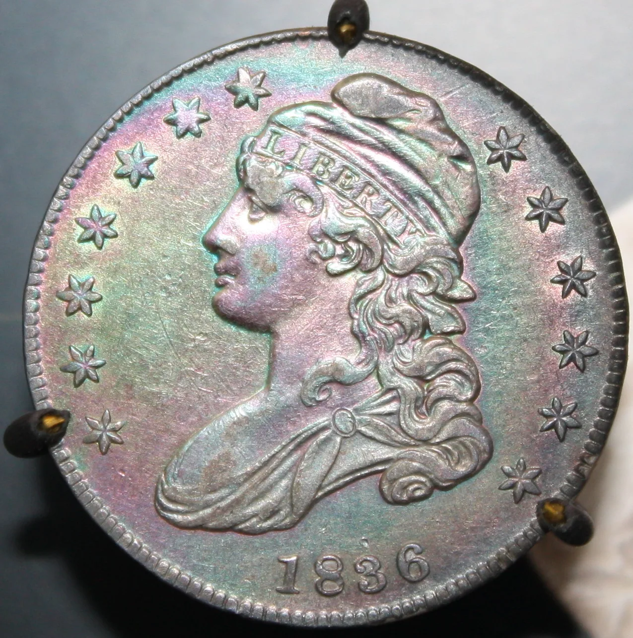 Obverse of an 1836 Capped Bust half dollar with rainbow toning, held in a display mount whose prongs cover part of the edge