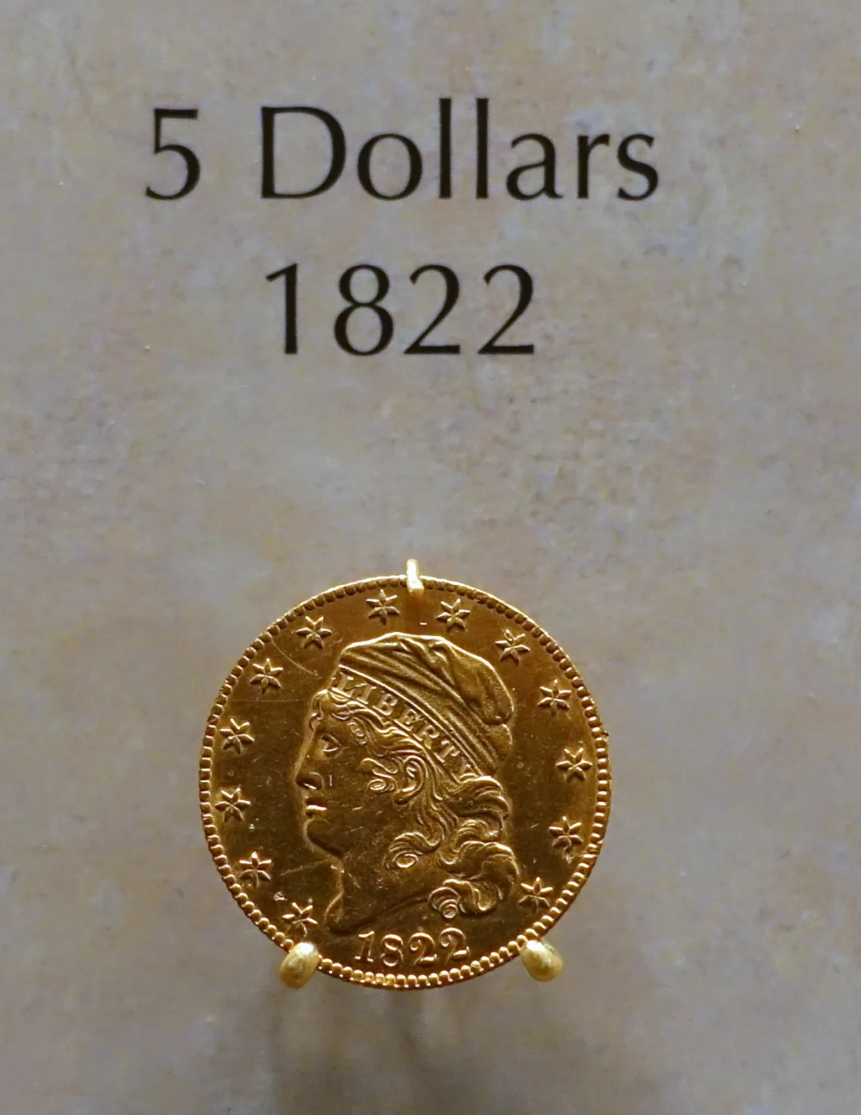 An 1822 half eagle mounted in display clips on a stone panel, below a printed placard reading 5 Dollars 1822