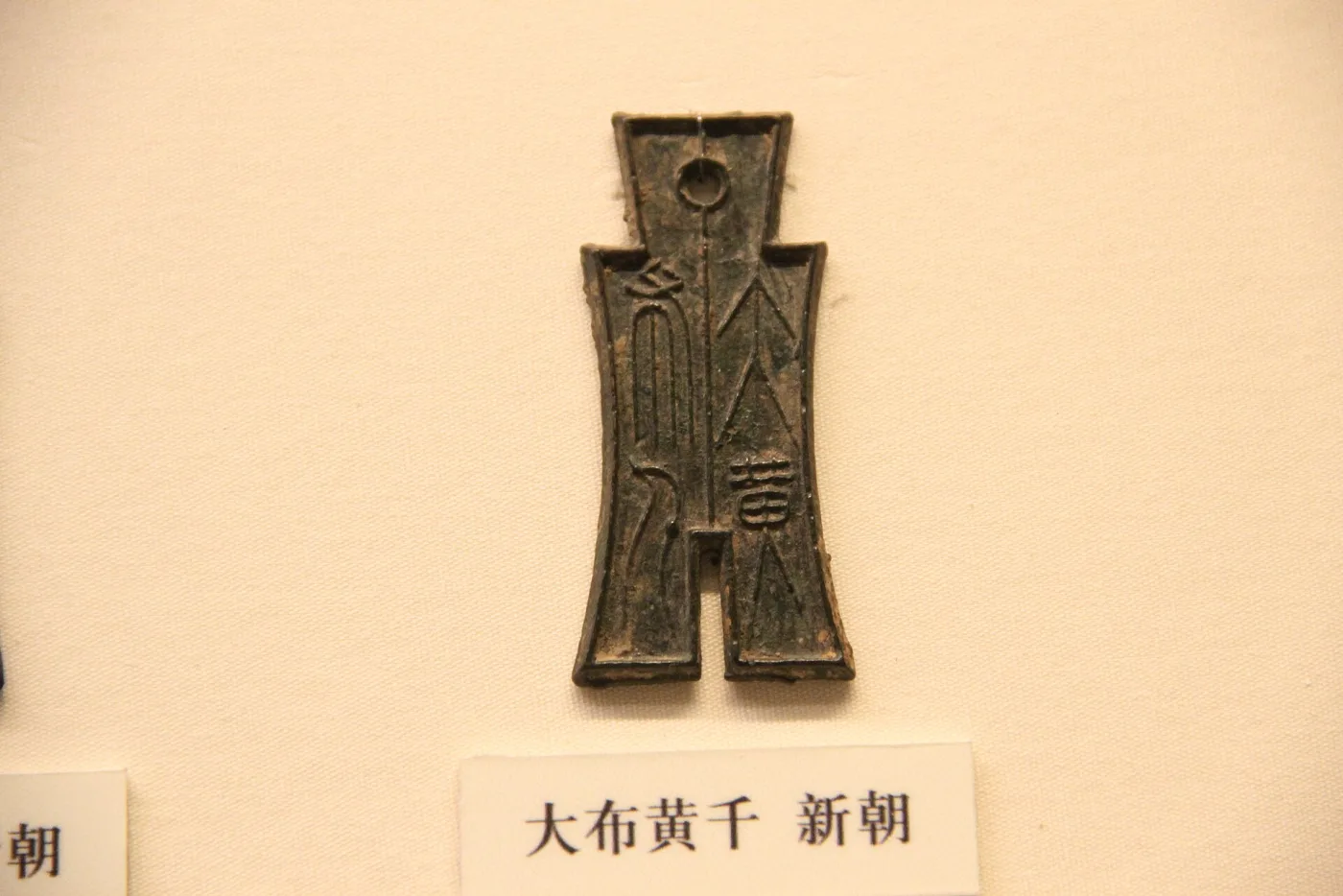 A bronze coin shaped like a small spade with two pointed feet and a squared handle, inscribed with vertical characters, displayed on a museum mount