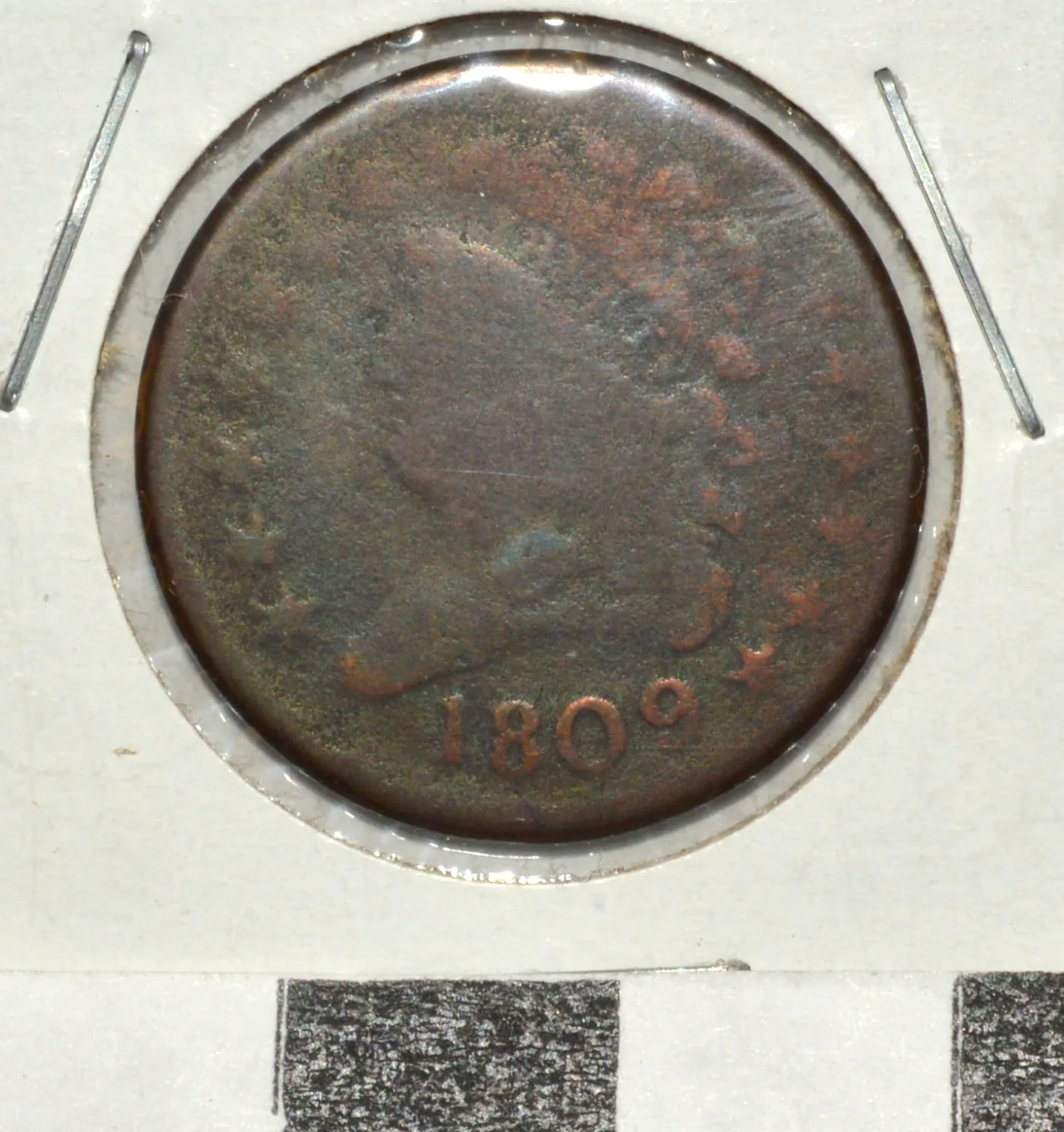 Obverse of a heavily worn 1809 Classic Head half cent in a paper coin holder, the portrait reduced to a smooth outline with the date 1809 still readable at the bottom