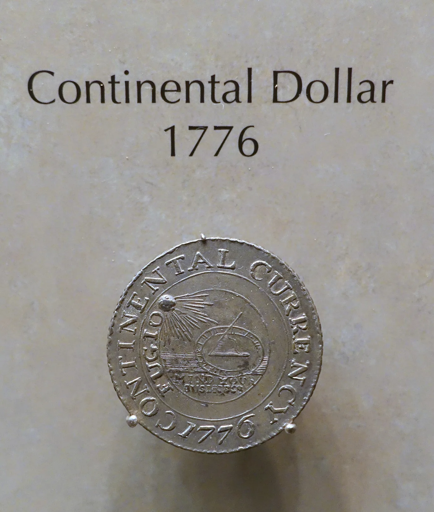 A pewter 1776 Continental dollar on museum display, showing the sundial beneath a radiant sun with FUGIO and MIND YOUR BUSINESS inside the legend CONTINENTAL CURRENCY 1776