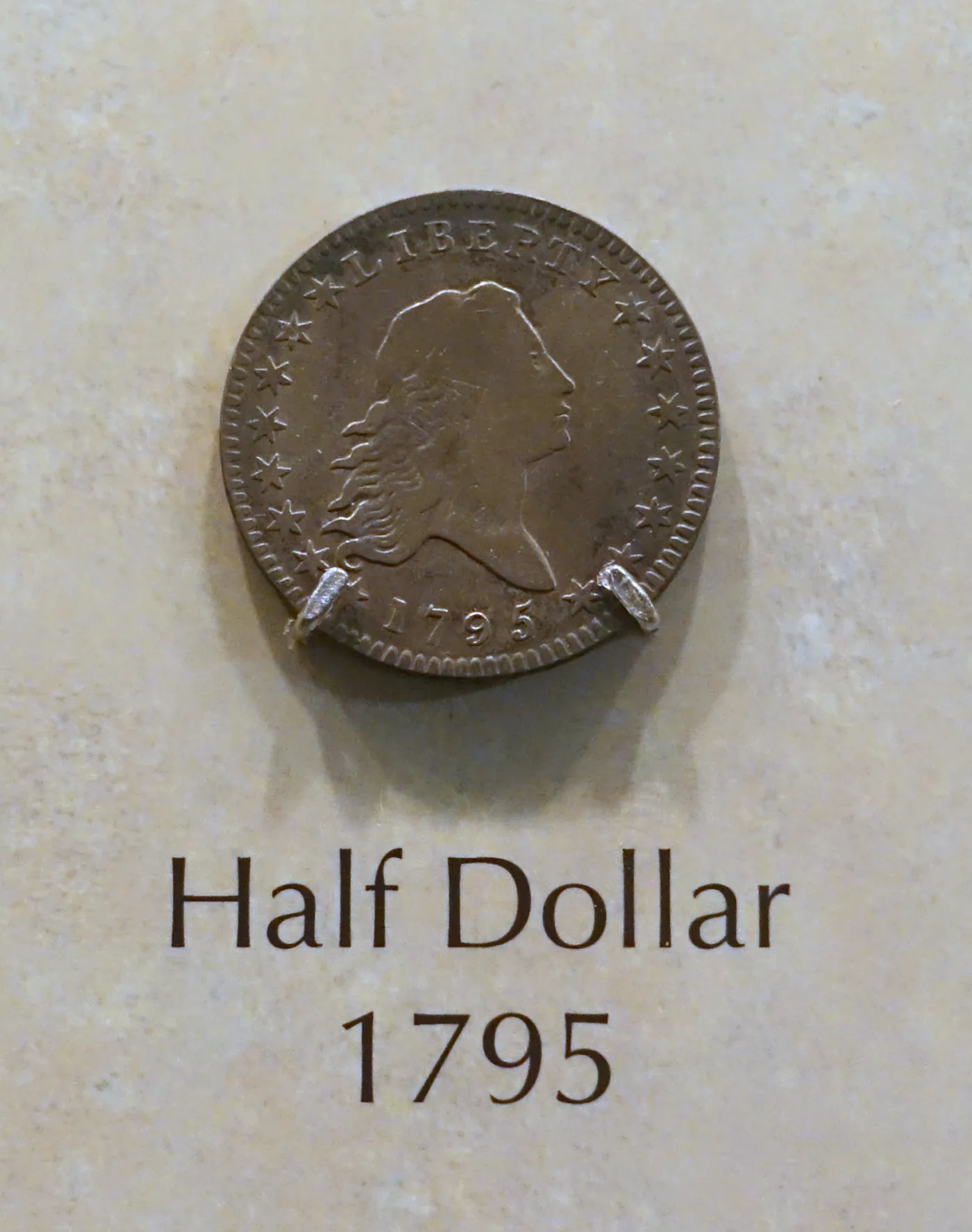 A 1795 Flowing Hair half dollar mounted on prongs in a museum display case, its obverse facing out beside a printed label reading Half Dollar 1795