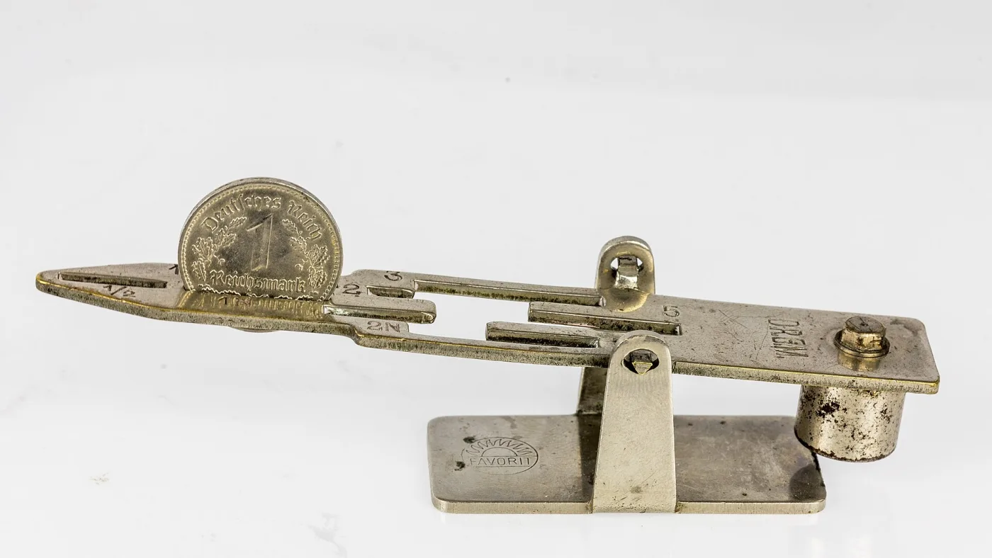 A small nickel-plated balance gauge marked with denominations, a German 1 Reichsmark coin standing in one of its slots