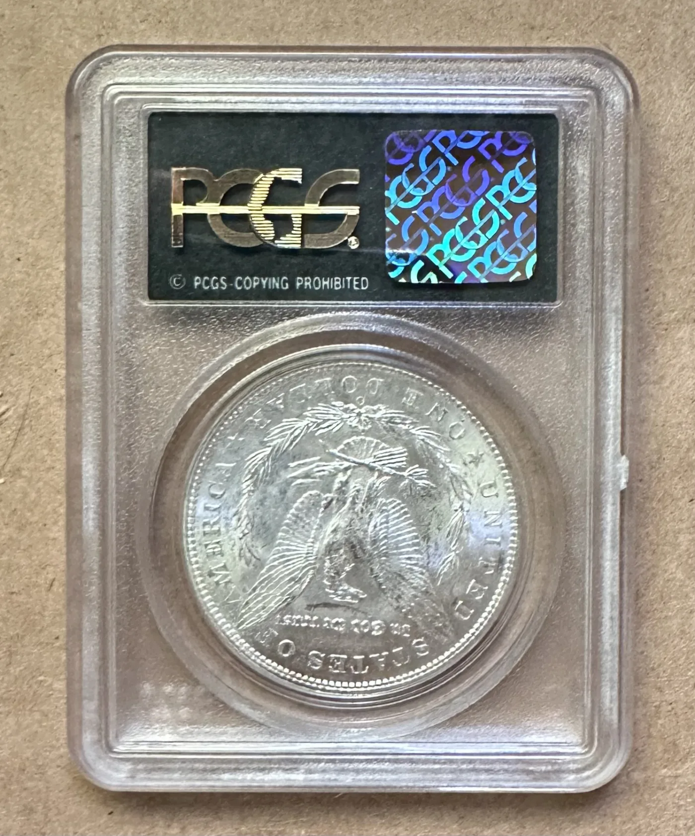 Back of a PCGS holder showing a security hologram beside the service logo, with the reverse of the encapsulated Morgan dollar visible below