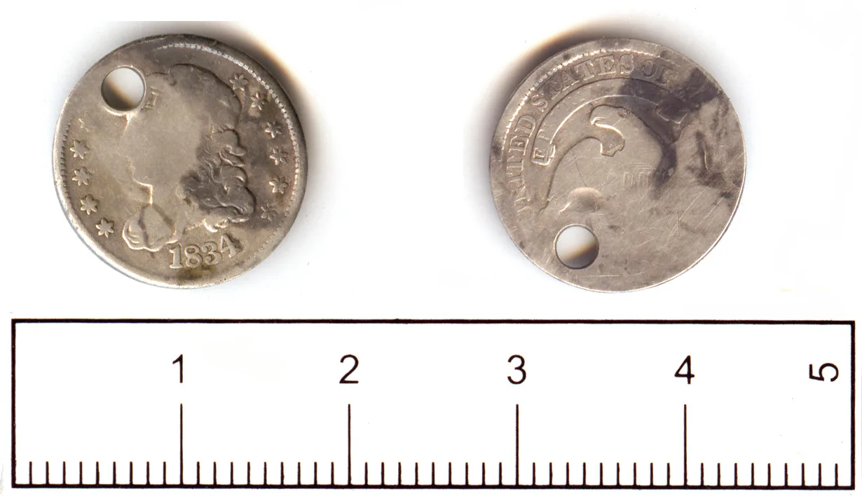Obverse and reverse of an 1834 United States half dime pierced by a hole near the rim, photographed beside a centimetre scale