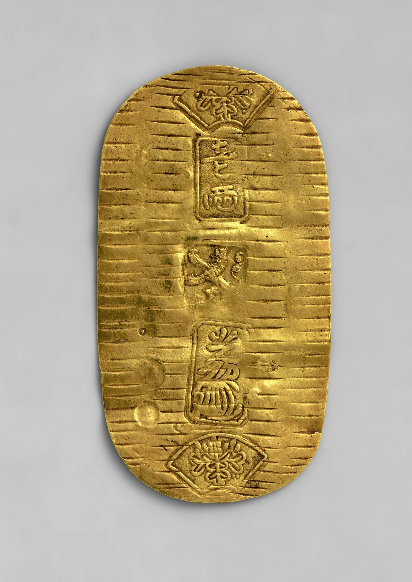Japanese gold koban covered in fine parallel file lines, with fan-shaped stamps at top and bottom and a small shield-shaped countermark in the centre