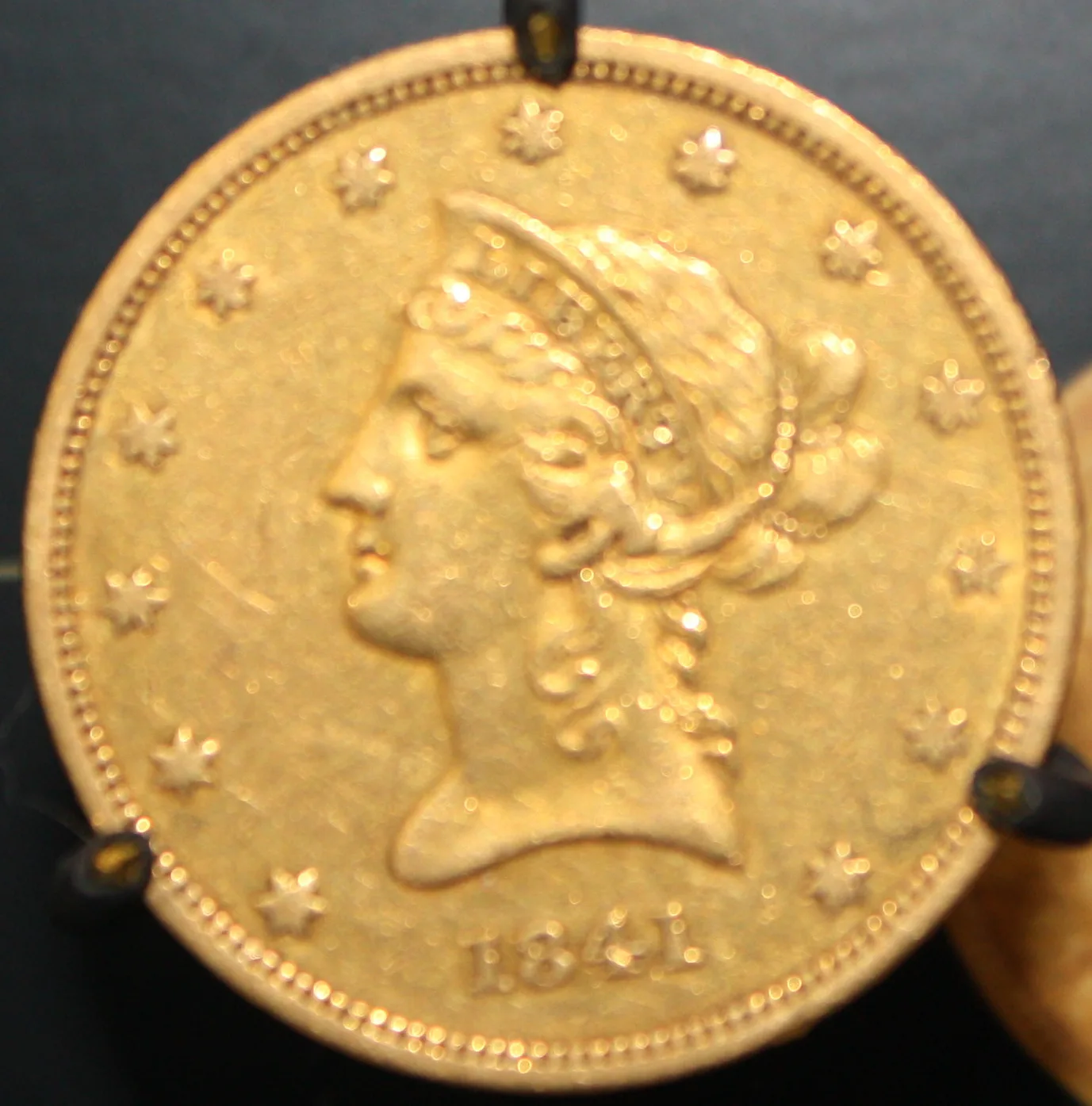 An 1841 Liberty Head Eagle gripped by four metal prongs of a jewelry bezel that press into the rim and the field