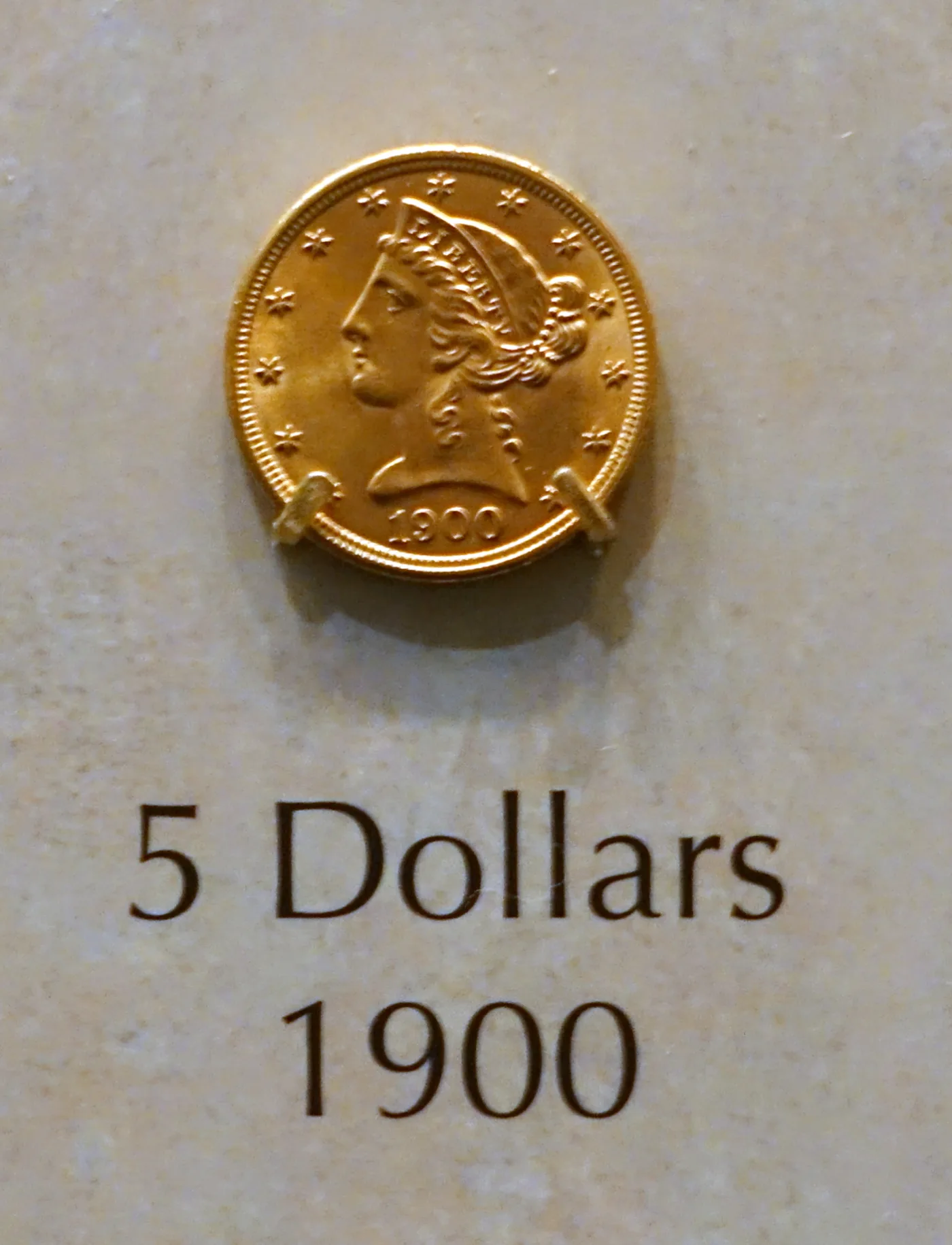 A 1900 Liberty Head Half Eagle mounted in a museum case beside a printed label reading 5 Dollars 1900