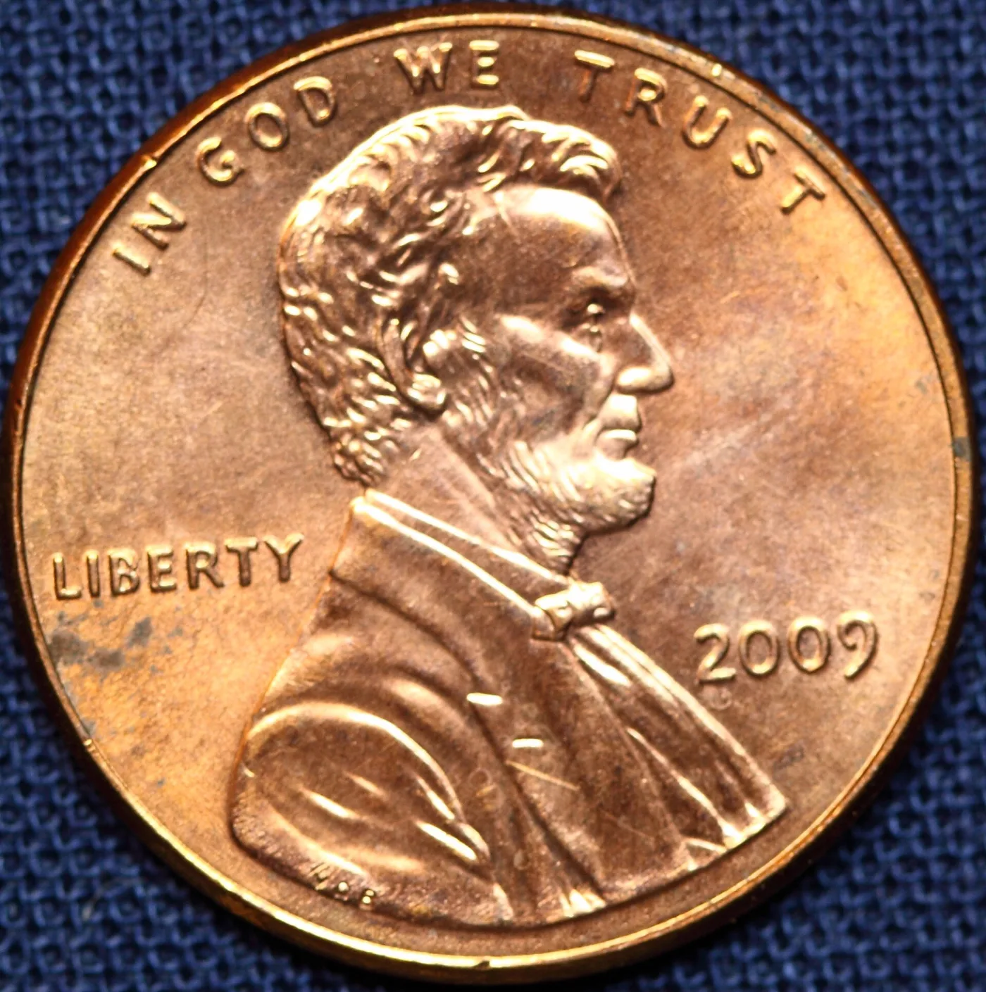 A circulated 2009 Lincoln cent lying on blue cloth, its copper plating unevenly toned with dull patches