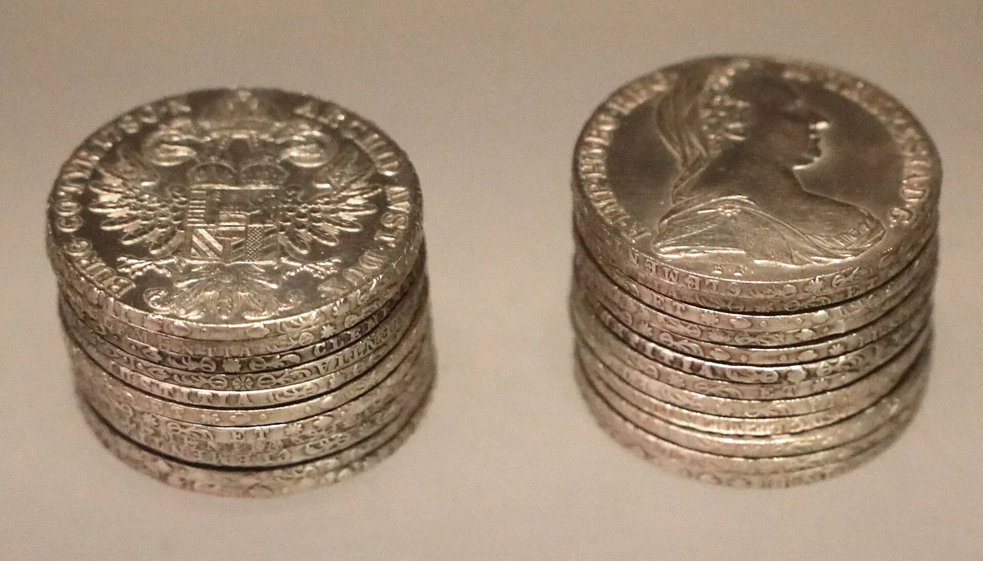 Two stacks of Maria Theresa thalers in a museum case, one showing the eagle reverse and one the portrait, with lettered edges visible down the sides
