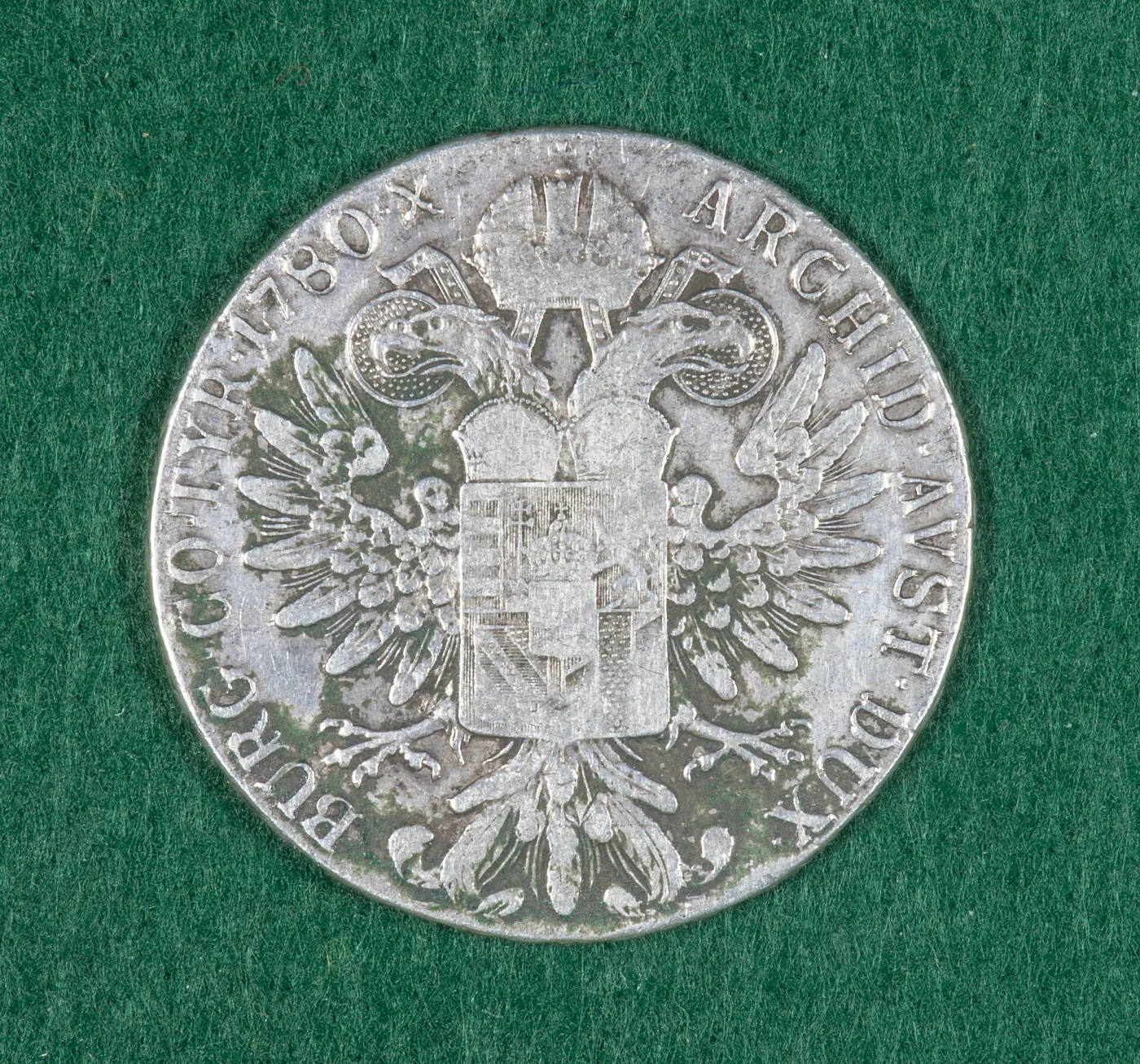 Heavily worn reverse of a 1780 Maria Theresa thaler on green felt, with the eagle and shield flattened and scratched