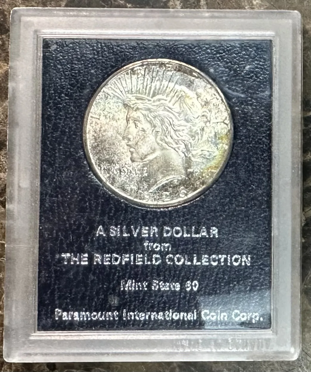 A toned Peace dollar mounted in a rigid black-and-clear Paramount International plastic holder marked A Silver Dollar from the Redfield Collection, Mint State 60