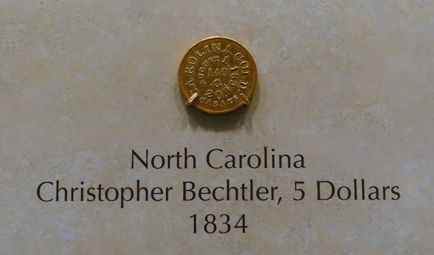 An 1834 Christopher Bechtler five dollar gold piece on museum display, lettered CAROLINA GOLD around AUGUST 1 1834, 140 G. and 20 CARATS, above a label reading North Carolina, Christopher Bechtler, 5 Dollars, 1834
