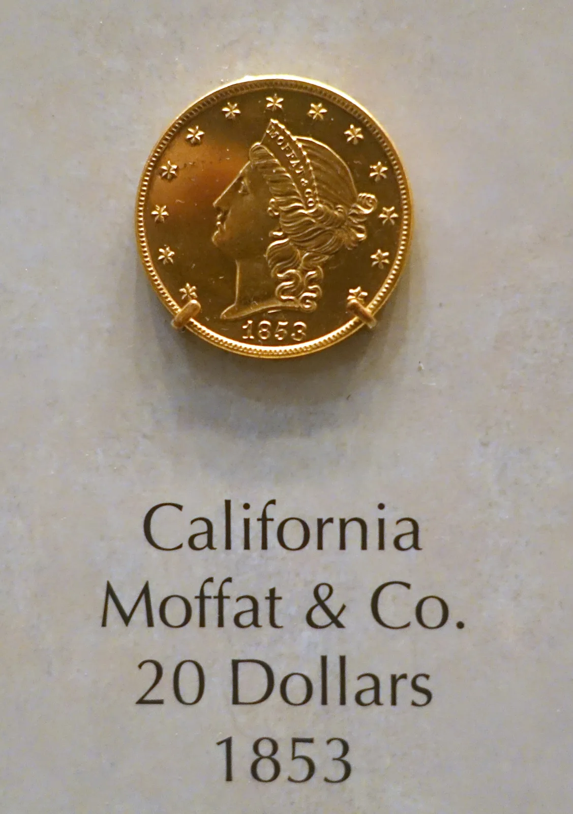 An 1853 Moffat and Company twenty dollar gold coin on museum display, a Liberty head inside thirteen stars with MOFFAT and CO. on the coronet and the date 1853, above a label reading California, Moffat and Co., 20 Dollars, 1853