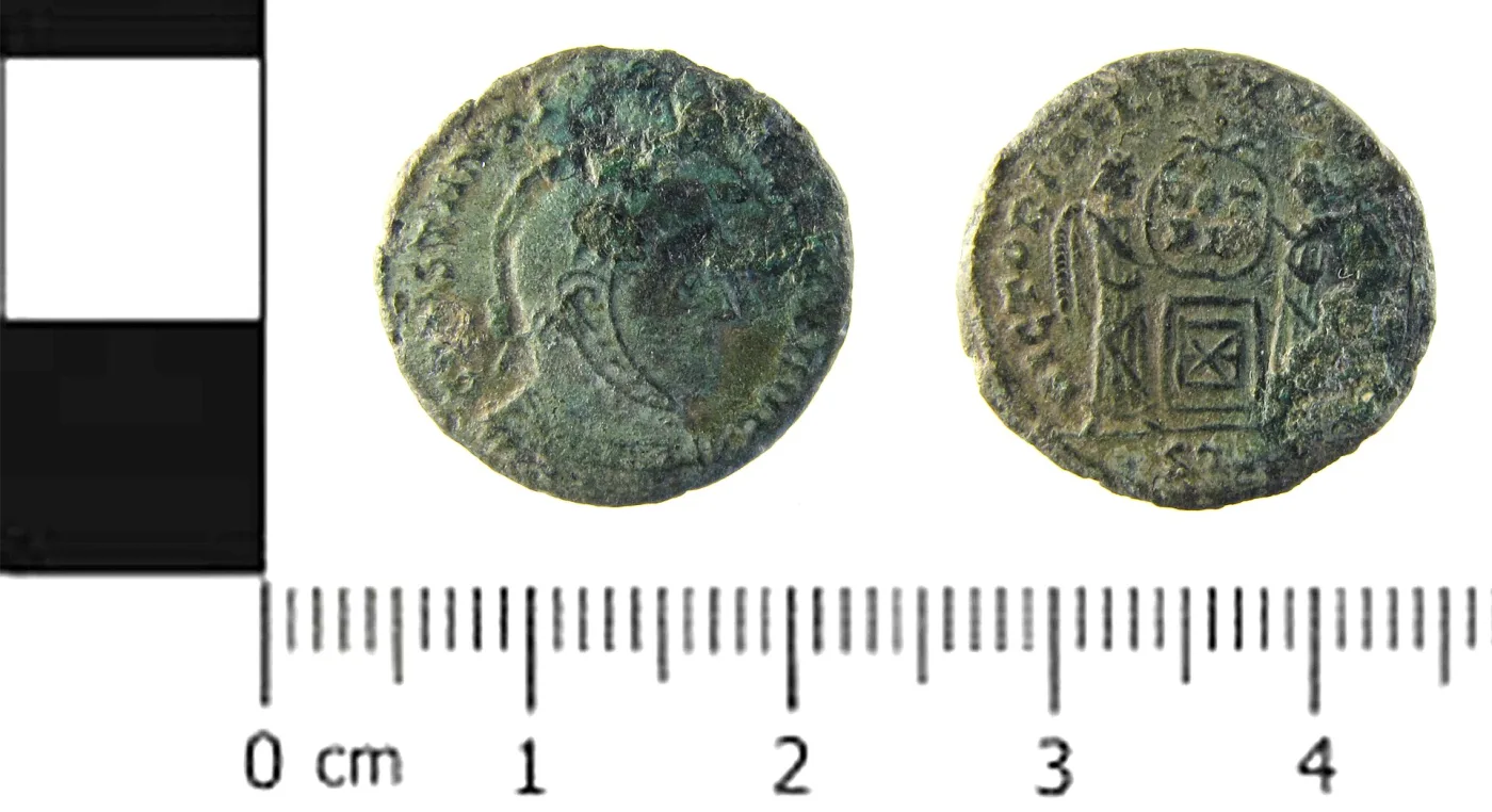 Obverse and reverse of a Roman copper-alloy nummus of Constantine I beside a centimetre scale: a helmeted bust on the front, and on the back two Victories holding a shield over an altar
