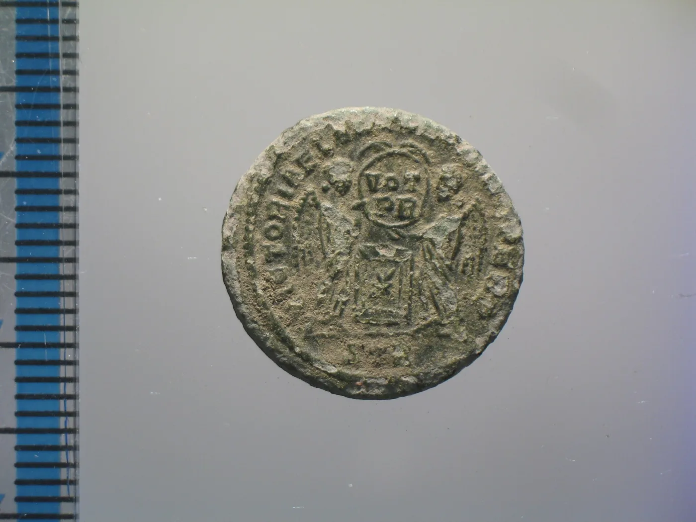 Reverse of a Roman nummus of Constantine I showing two winged Victories holding a round shield inscribed VOT PR above an altar, beside a millimetre scale