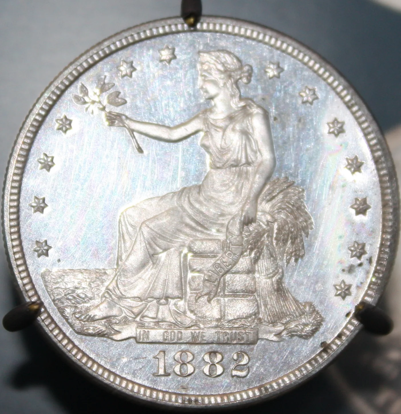 Obverse of an 1882 proof Trade Dollar held by mount prongs, showing brilliant reflective fields against frosted design elements